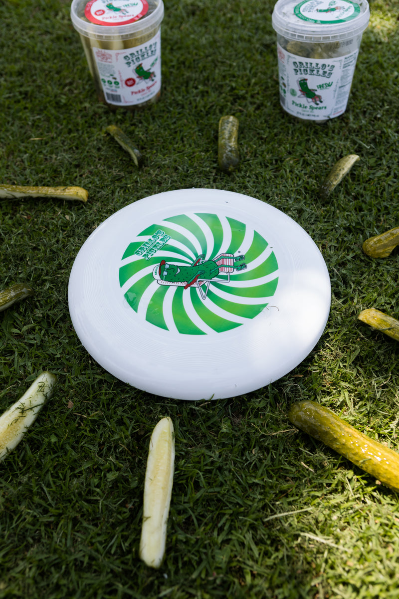 frisbee with pickle on lawn chair and green swirl background laying on grass with pickles surrounding it 